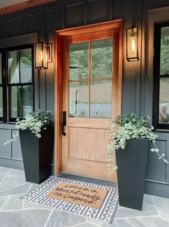 a modern porch with a stained door, black tall planters with greenery and layered rugs looks elegant and laconic and isn't difficult to recreate