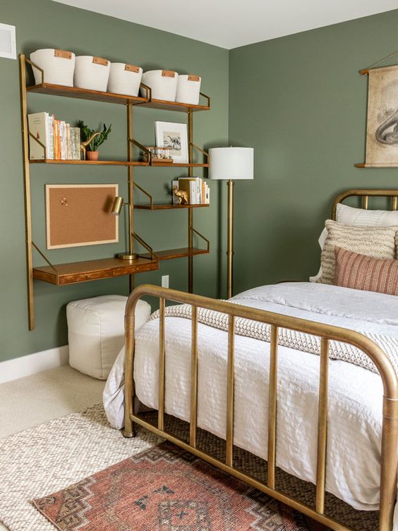 a modern vintage bedroom with green walls, a brass bed with neutral bedding, a shelving unit with a tiny desk