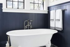 a navy bathroom clad with beadboards, with grey tiles, a white tub in vintage style and a couple of windows