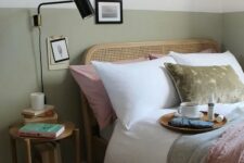 a pretty and welcoming bedroom with white and olive green walls, a rattan bed and neutral and green bedding, a wooden nightstand and a black sconce