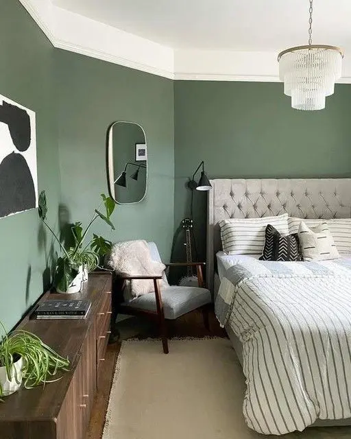 a sage green bedroom with a grey upholstered bed and printed bedding, dark-stained furniture, some chic decor and plants