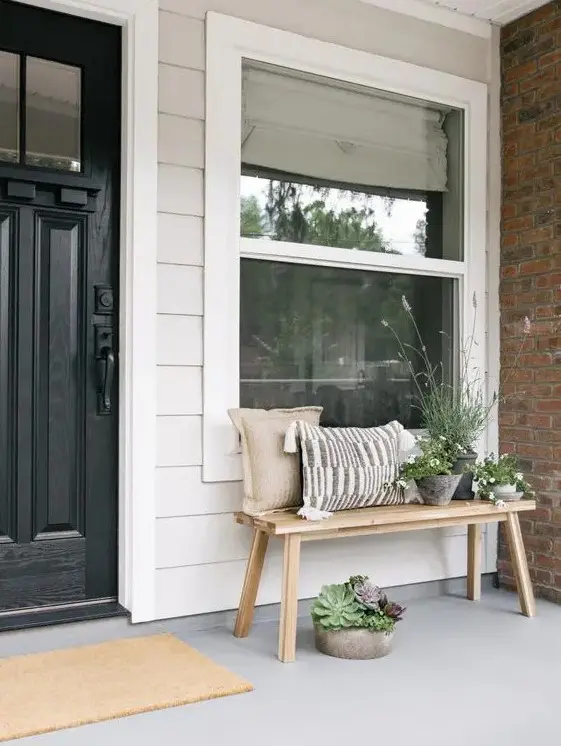 a simple modern front porch with a wooden bench and pillows, some potted greenery and succulents and a black door