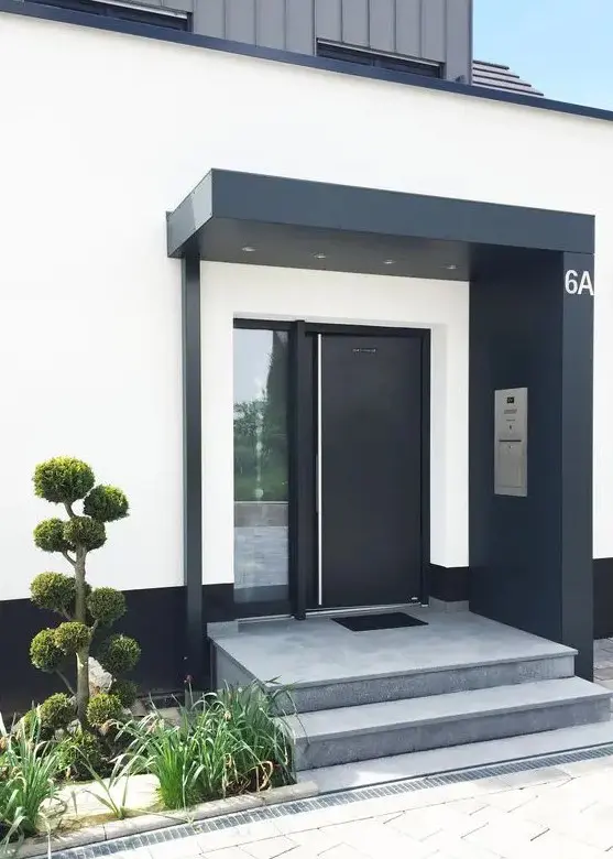 a super chic modern entrance with a concrete ladder, a black over with lights, a black door and greenery next to the ladder