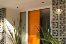 a very chic mid-century modern front porch with a bold orange door and planters with oversized plants plus screens to protect from the sun