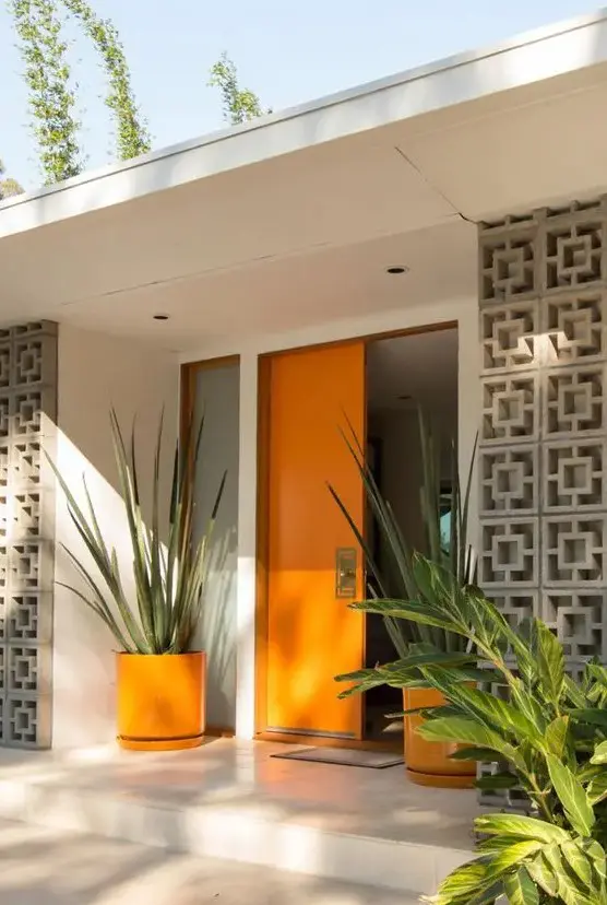 a very chic mid-century modern front porch with a bold orange door and planters with oversized plants plus screens to protect from the sun