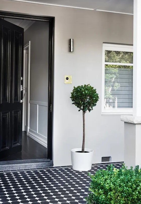 a very laconic and simple modern porch with a black door, black and white patterned tiles, some potted greenery is a chic idea to repeat