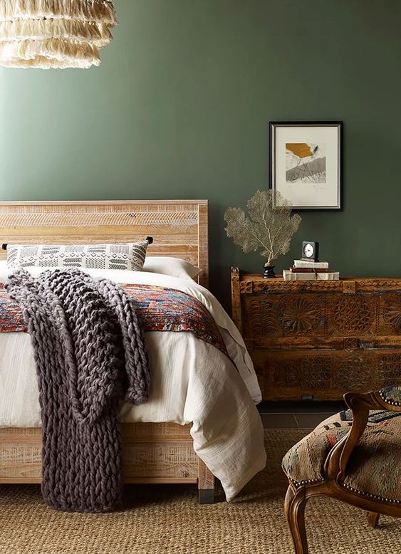 a vintage bedroom with green walls, a rough wooden bed with printed bedding, a dark-stained dresser, a chair and a jute rug