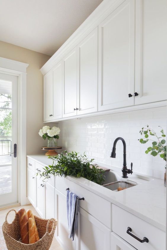 a welcoming white kitchen with white subway tiles, a white countertop, black fixtures and some blooms and greenery