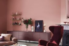 an exquisite living room with pink walls, a burgundy velvet chair and a burgundy credenza, a brown sofa and a fireplace