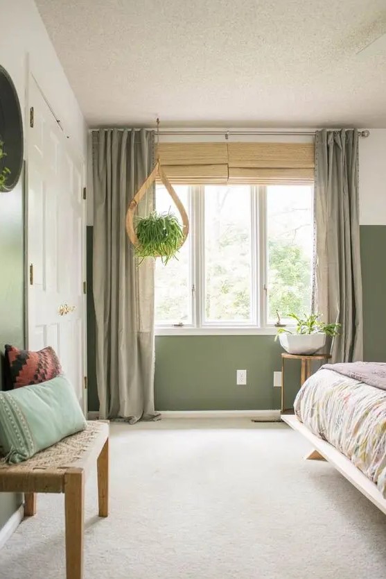 an olive green boho bedroom with a bed with printed bedding, a woven bench and pillows, a potted plant and lovely curtains