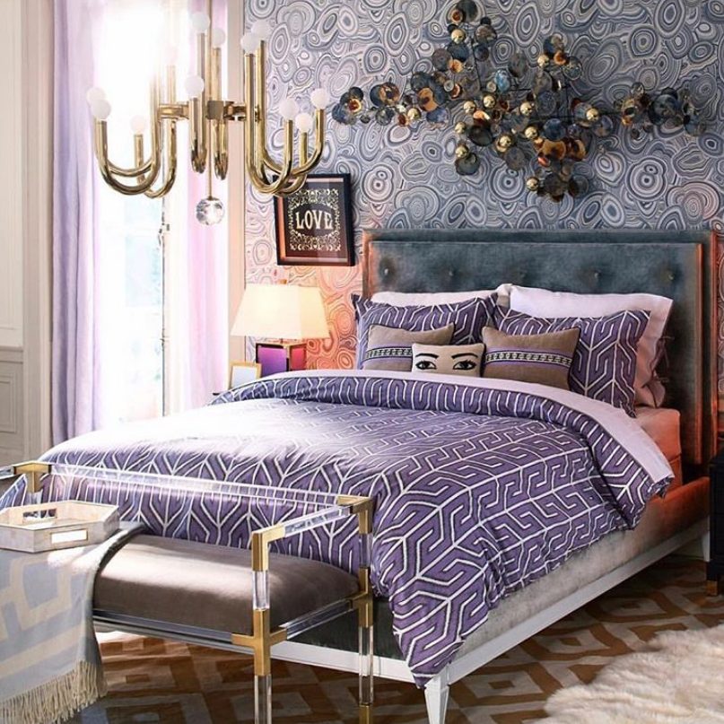 A maximalist bedroom with a printed accent wall, a bold artwork, a glam chandelier and a bench with gold touches and lilac and purple linens
