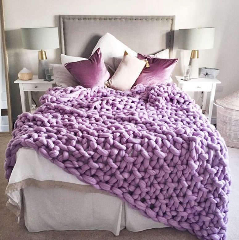 a white and grey bedroom with elegant and simple furniture and lamps and gorgeous purple pillows and a chunky knit blanket