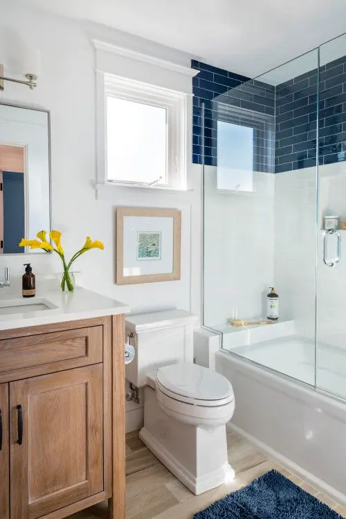 a beach bathroom with white and navy tiles, a tub with a shower space, a timber vanity, artwork and a navy rug