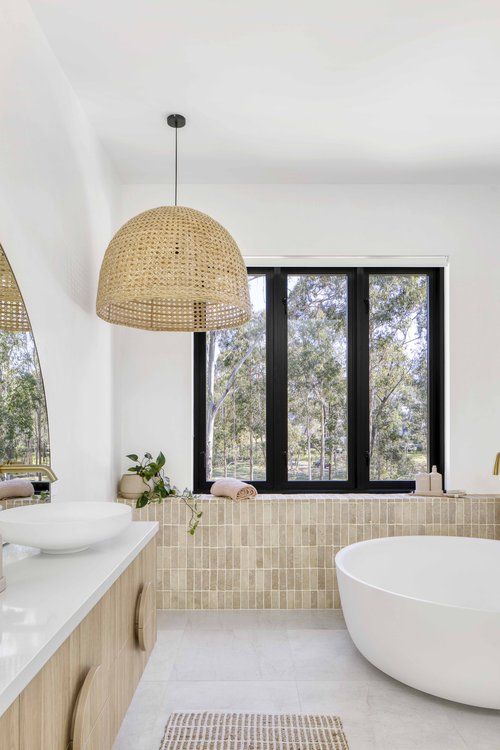 a beach bathroom with white walls and tan tiles, a round tub, a timber vanity and roudn sinks, a woven pendant lamp and a window with a view