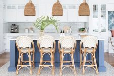 a beach kitchen with white cabinetry, a navy kitchen island, rattan chairs, woven lamps and greenery