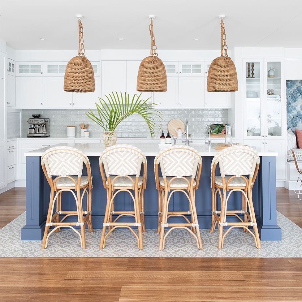 A beach kitchen with white cabinetry, a navy kitchen island, rattan chairs, woven lamps and greenery