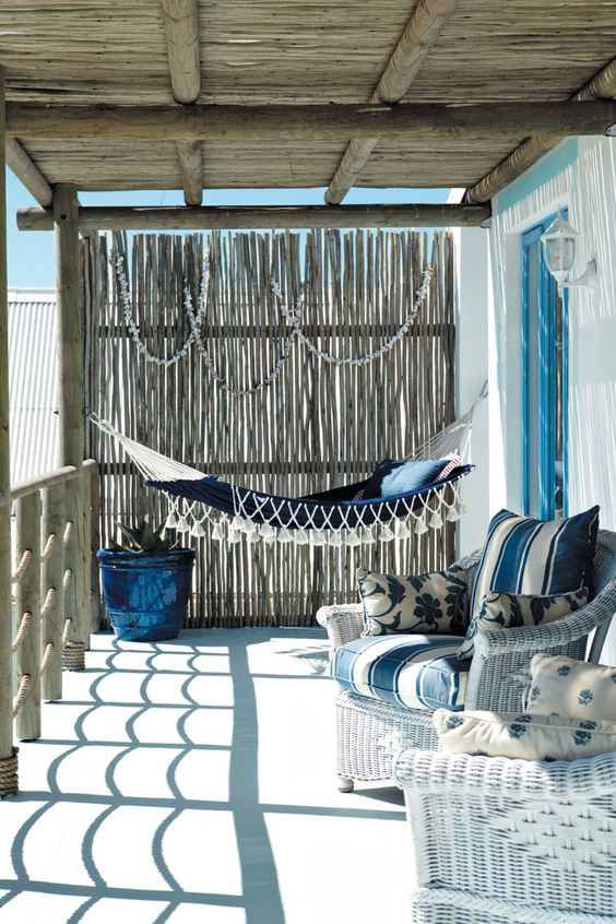 a beautiful beach porch with a navy hammock with tassels, white wicker chairs with blue upholstery and seashell garlands