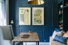 a blue home office with paneling on the walls and a stylish storage unit, neutral furniture, a white and gold lamp and a neutral chair