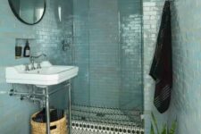 a boho coastal bathroom with blue skinny tiles, a boho printed tile floor, potted plants, a round mirror and black towels