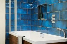 a bold seaside bathroom clad with large scale blue tiles, a wood clad tub and neutral faucets looks statement-like