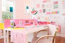 a bright and glam home office with a pink striped accent wall, a pegboard wall with lots of stuff, two desks and pink fringe