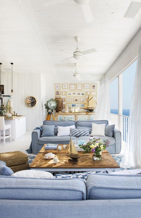 a bright and welcoming beach living room with a glazed wall, blue sofas, a wooden coffee table, a cool gallery wall and a beach sideboard