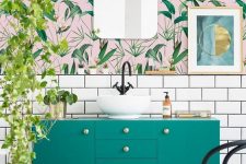 a bright bathroom with pink and green tropical wallpaper, an emerald vanity, cascading greenery and touches of gold
