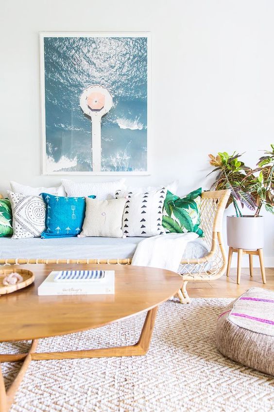 a bright coastal living room with a rattan sofa, colorful pillows, a woven rug and ottoman and a beach artwork