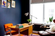 a bright mid-century modern home office with navy walls, a rich stained desk, an orange chair, a bright rug and a leather lounger