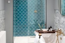a bright seaside bathroom with turquoise fishscale tiles, a vintage clawfoot tub, a wooden mat and touches of gold