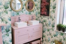 a bright tropical bathroom with tropical wallpaper, a blush vanity with stools, gold framed mirrors and a neon sign