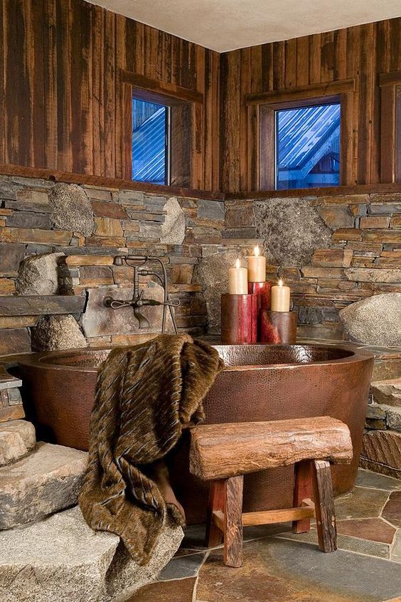 a chalet bathroom clad with stone and wood, with a statement copper tub, rough rocks and a wooden stool