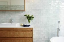 a charming coastal bathroom clad with very pale green glazed tiles, a tub, a wooden vanity and a pendant lamp