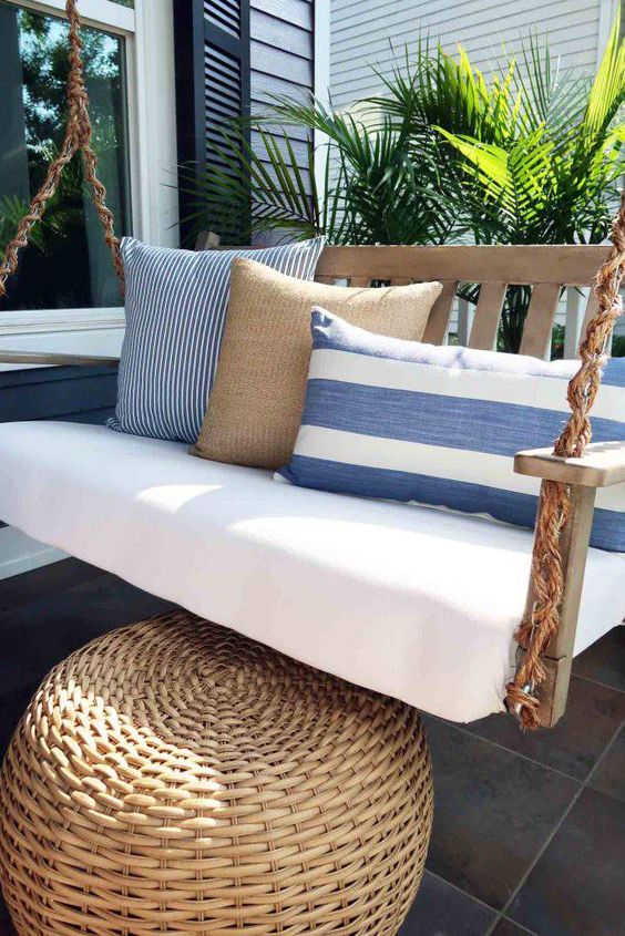 a chic coastal front porch with a hanging upholstered daybed with blue pillows and a wicker ottoman plus potted plants