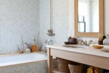 a coastal bathroom clad with small scale blue tiles, a tub, a wooden vanity, baskets and seaside decor and a mirror in a wooden frame