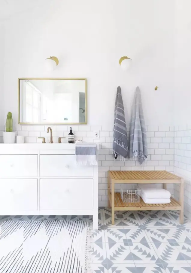 a coastal bathroom done with white subway and blue graphic tiles, a white vanity, a wooden bench, some towels and potted plants