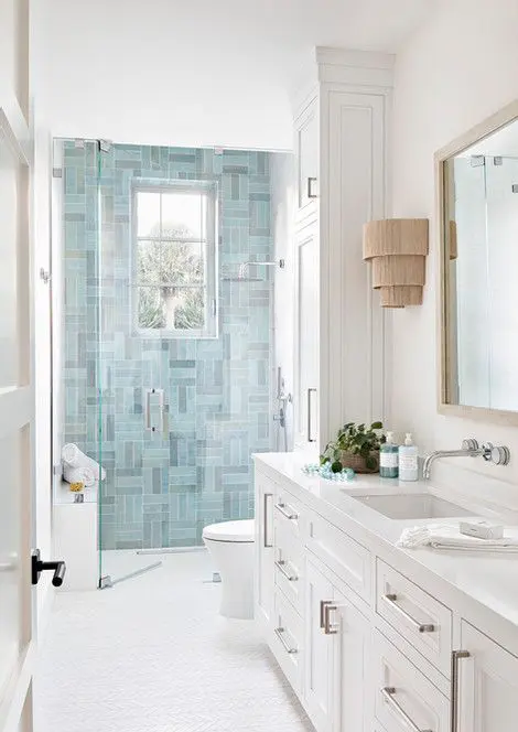 a coastal bathroom with blue tiles in the shower, a white vanity and white tiles, a mirror and some wall sconces