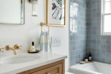 a coastal bathroom with white walls, blue Zellige tiles around the tub, penny tiles on the floor, a timber vanity and some art