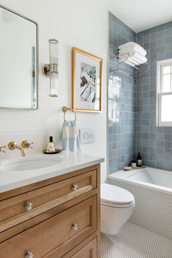 a coastal bathroom with white walls, blue Zellige tiles around the tub, penny tiles on the floor, a timber vanity and some art