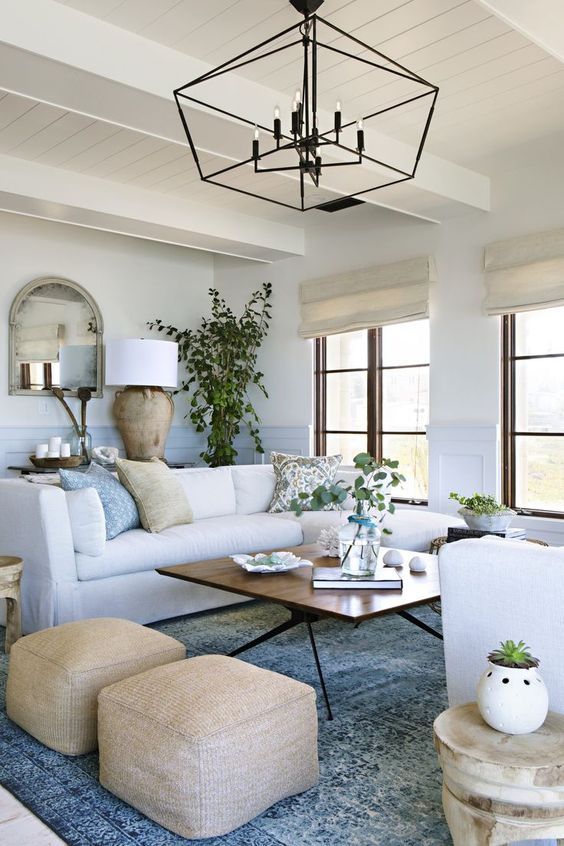 a coastal farmhouse living room in white, with light blue paneling, a chandelier, potted greenery, candles and tan ottomans