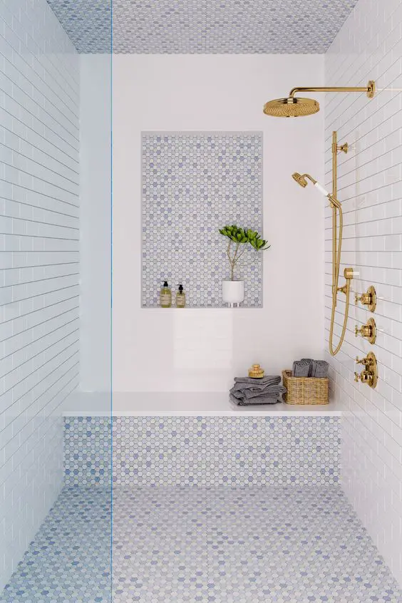 a coastal shower space done with white and blue tiles, a niche, a bench, some towels and gold fixtures