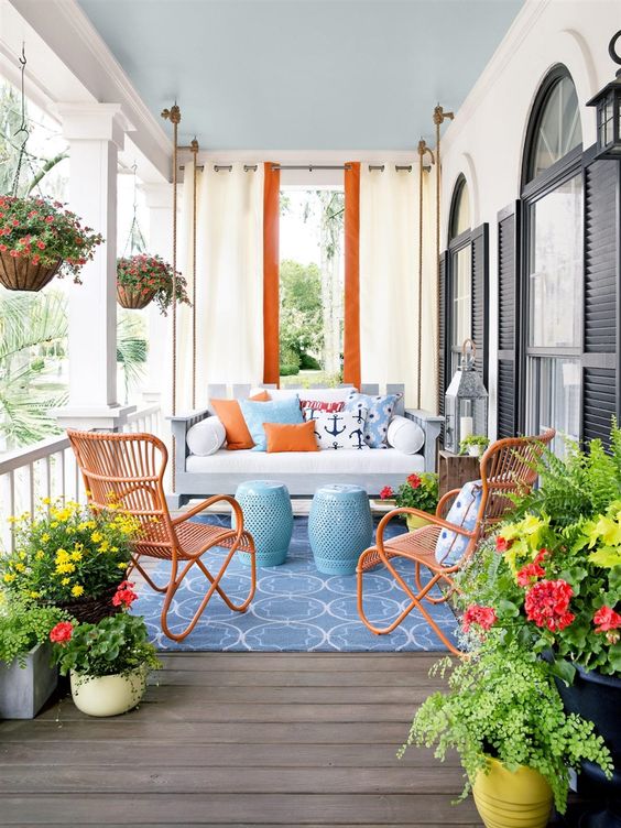 a colorful nautical porch with a suspended daybed with colorful printed pillows, orange rattan chairs, candle lanterns and potted blooms