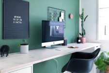 a contemporary home office with a hunter green statement wall, a neutral desk, a gallery wall and potted plants