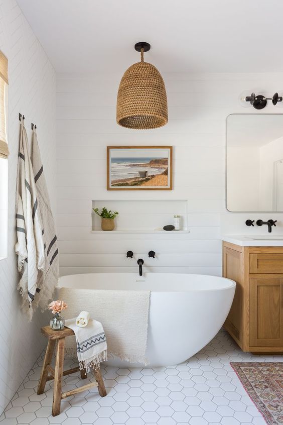 a cool beach bathroom with white shiplap walls, a white hex tile floor, a timber vanity, an oval tub, some artwork, black fixtures and a woven pendant lamp