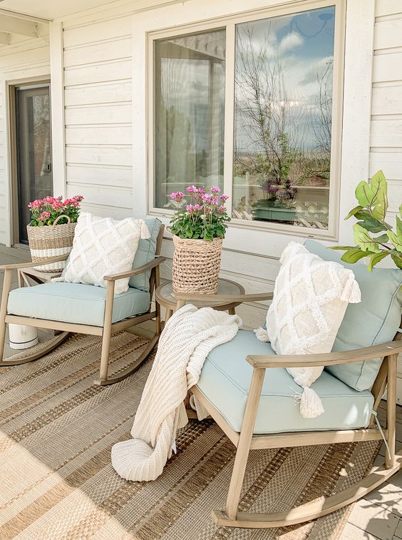 a cozy coastal porch with a jute woven rug, wooden chairs with pale blue upholstery, potted blooms and tassel pillows