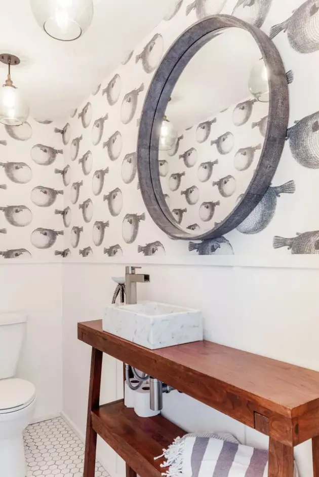 a creative coastal bathroom done with fish print wallpaper, a round mirror, a wooden vanity with a small sink and some lamps