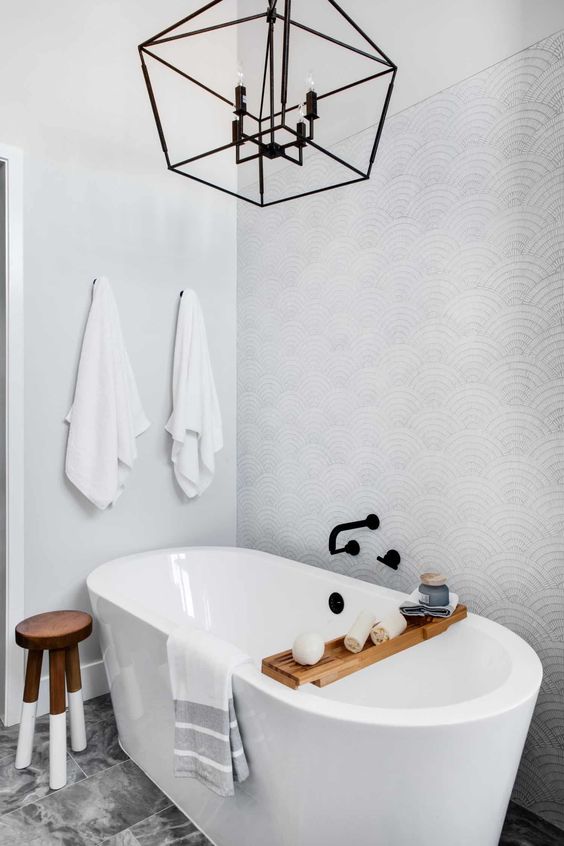 a delicate coastal bathroom done with wallpaper, an oval tub, a tiled floor, a frame pendant lamp and some wooden touches