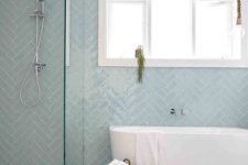 a fresh coastal bathroom with a wall clad with light blue herringbone tiles, a free-standing tub, a wooden stool and a basket