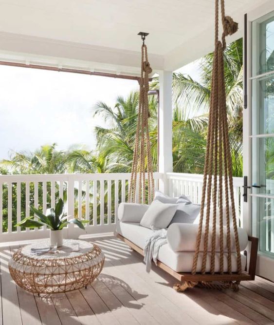 a lovely coastal porch with a suspended upholstered daybed, a rattan coffee table and neutral pillows is welcoming