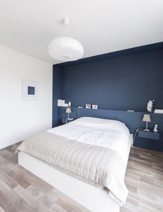 a minimalist bedroom with a navy accent wall and floating nightstands, a white bed and a pendant lamp plus an artwork